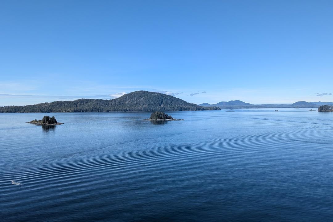 Calm blue waters and small forested islands seen from a cruise ship balcony under a clear sky during an Inside Passage transit.