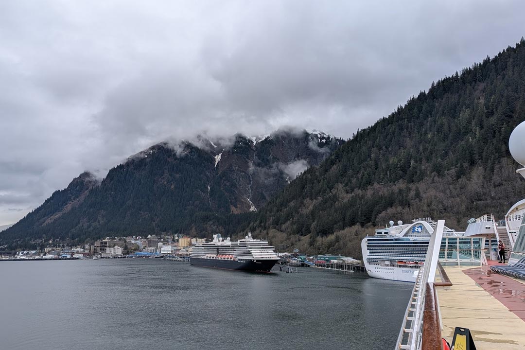 View looking down the side promenade deck of a cruise ship docked in Juneau, Alaska, with steep fog-covered mountains rising directly behind the port.