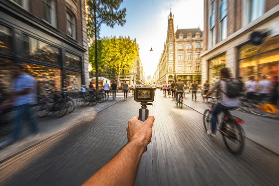 A traveler holding an action camera while walking through a lively city street with motion blurred at the edges.