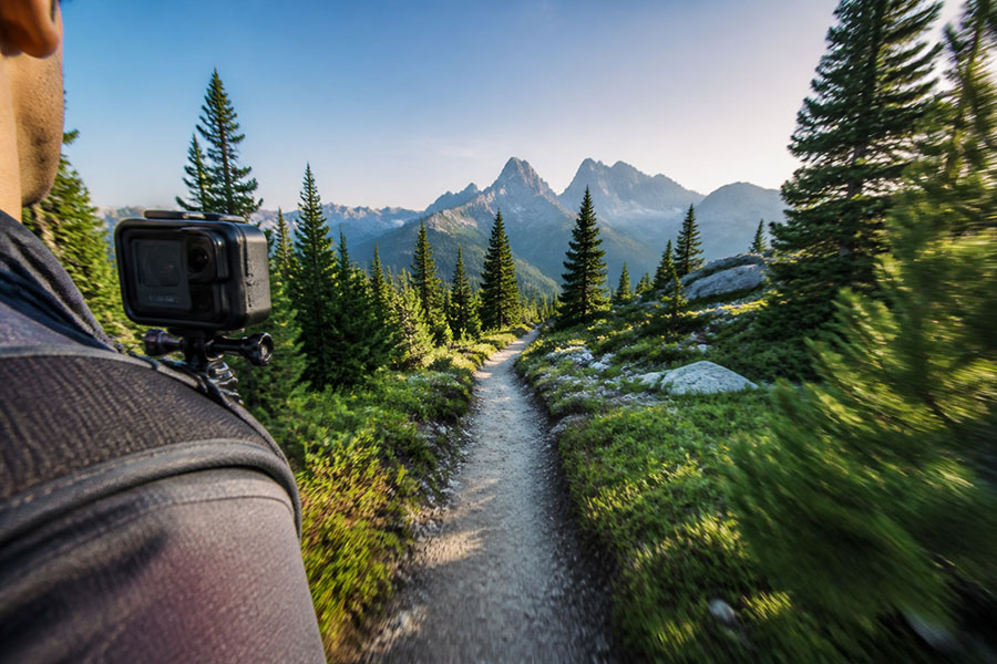 A hiker wearing an action camera on a backpack strap while moving along an alpine trail.